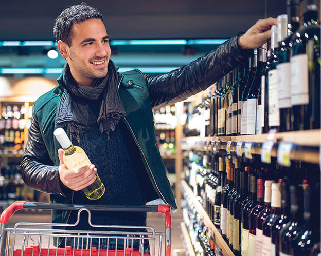 Young man with a shopping cart is in a supermarket picking up wine 